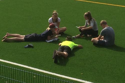 Rippikoulun raamisryhmä meneillään isosen johdolla. Nuoria istumassa ja makaamassa vihreällä jalkapallokentällä.