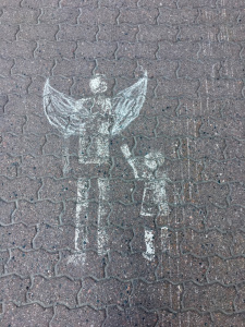 Kuva katumaalauksesta, jossa kävelytien merkin aikuiselle on piirretty enkelin siivet ja käsi, joka pitää lapsesta kiinni.