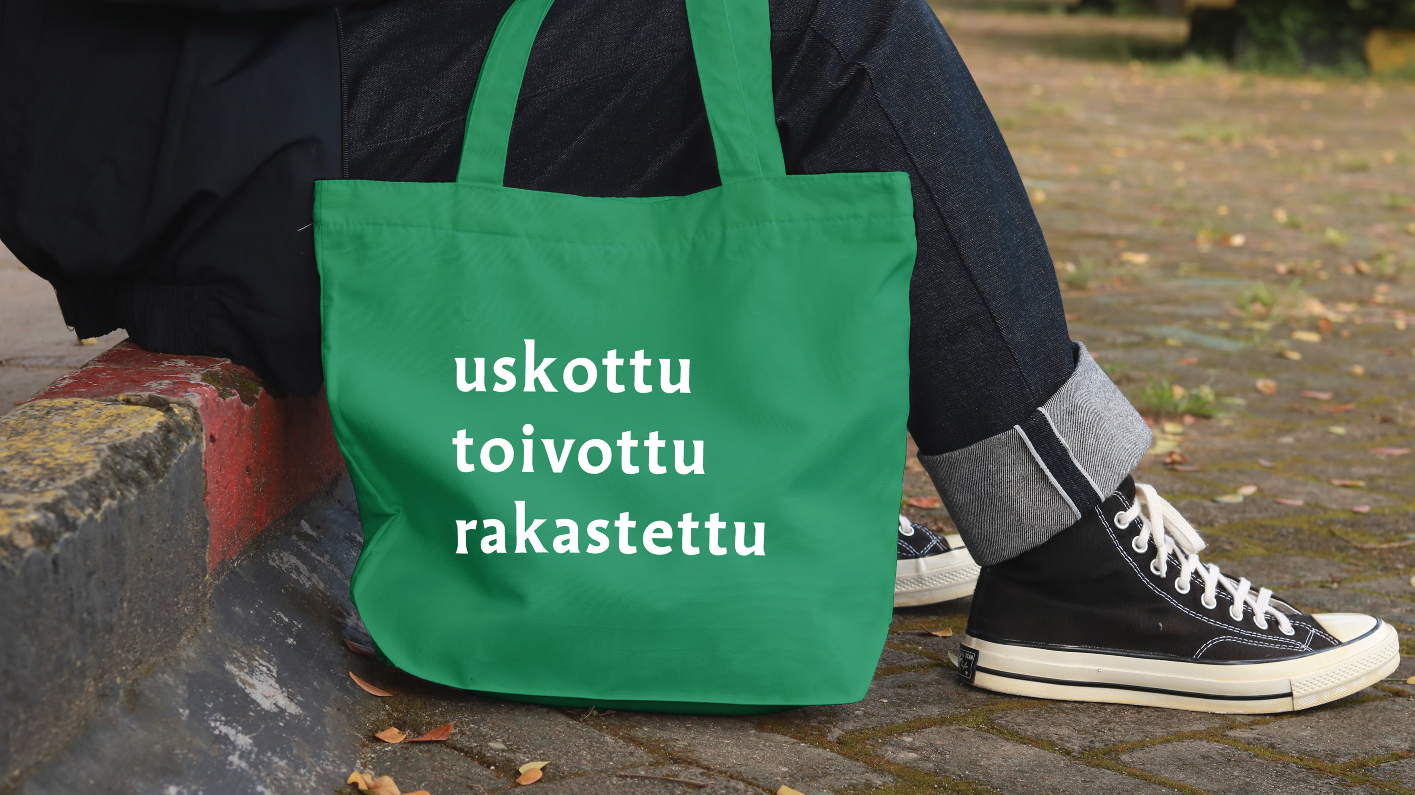 Katukiveyksellä istuvan henkilön jalat, etualalla kangaskassi, kyljessä lukee uskottu toivottu rakastettu